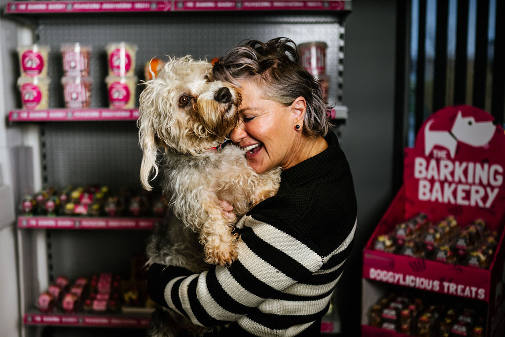 Woman holding a small dog in front of a store shelf with 'The Barking Bakery' products.